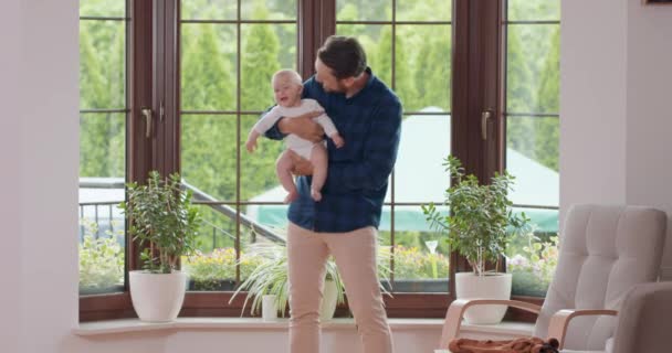 O pai barbudo e com bigode segura seu bebê adorável e acena levemente nas pernas, sorrindo na frente da janela, depois se vira para a janela Janela e uma parede branca no fundo — Vídeo de Stock
