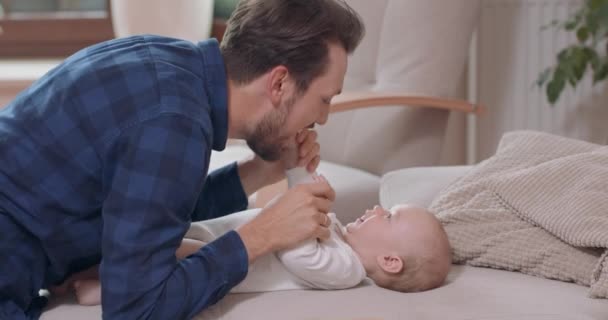 Sakallı ve bıyıklı baba, bebek kanepede diz çöküyor, bebek yüzüne nazikçe hava üflüyor bebek başını sallıyor ve babasının çenesine elleriyle, kanepesiyle, yeşille dokunuyor. — Stok video