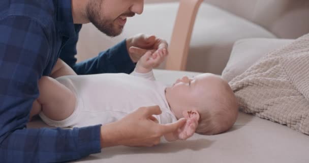 Sakallı ve bıyıklı babaya yaklaş, bebek kanepede yatıyor, elleriyle oynuyor, gülümseyen sandalye ve arka planda beyaz bir duvar var. — Stok video