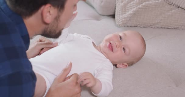 Primo piano del bambino sdraiato sulla schiena sul divano, sorridente e felice, guardando suo padre, e cattura le guance dei suoi padri, che si inginocchia accanto al bambino, gioca con esso sorride, solletica e parla. — Video Stock