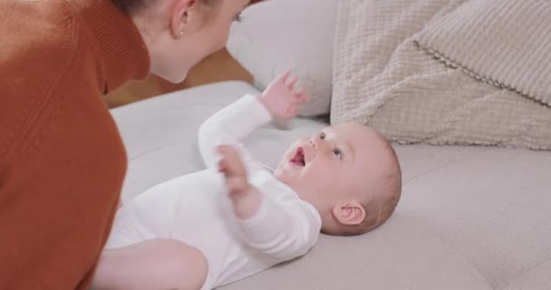 Acercamiento del bebé acostado boca arriba en el sofá, sonriente y feliz, estrechando las manos, mirando a su madre, que se arrodilla junto al bebé, habla con él y sonríe — Vídeos de Stock