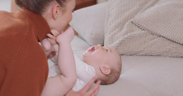 Κοντινό πλάνο του μωρού ξαπλωμένο ανάσκελα στον καναπέ, χαμογελαστό και χαρούμενο, εξεγερμένα χέρια και πόδια, κοιτάζοντας τη μητέρα του, που γονατίζει δίπλα στο μωρό, του μιλάει, του φιλάει και του χαμογελά. — Αρχείο Βίντεο