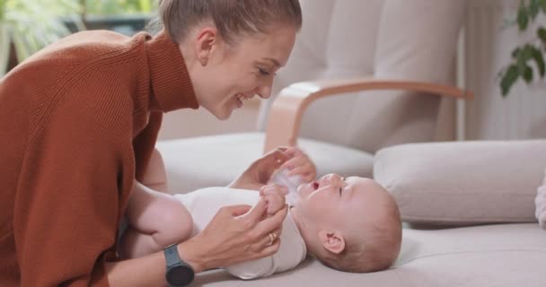 Primo piano del bambino sdraiato sulla schiena sul divano, sorridente e felice, mani e piedi sollevanti, guardando sua madre, che si inginocchia vicino al bambino, e sorride Sedia e una finestra sullo sfondo — Video Stock