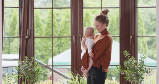 Mladá matka drží své roztomilé dítě a zůstává s ním před oknem, líbání dítě Okno, zelené rostliny, a bílá zeď na pozadí — Stock video