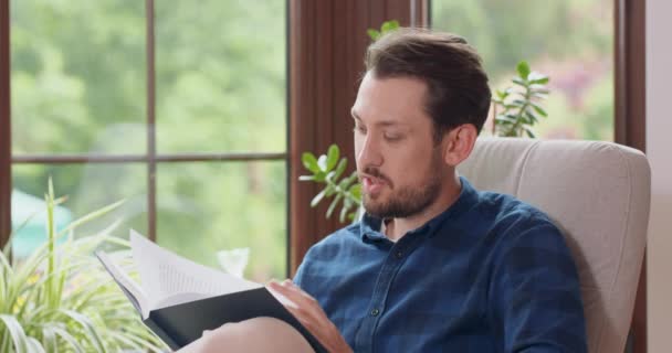 Крупним планом портрет молодого бородатого чоловіка, який читає книгу, сидячи в білому кріслі, молодий чоловік відпочиває вдома Вікно і зелені рослини на фоні — стокове відео