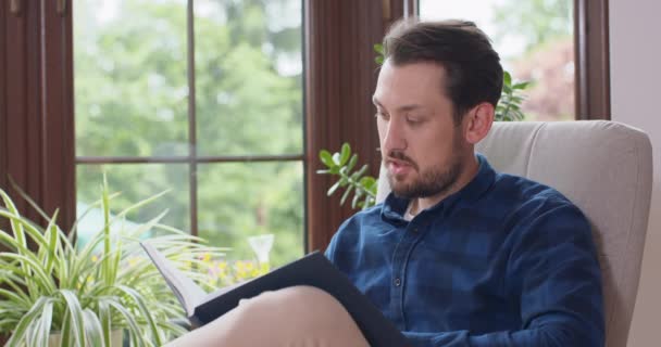 수염을 기른 젊은 남자가 흰 의자에 앉아 책을 읽고 있는 모습을 확대 해 보 세요. 젊은 남자가 집 창문에서 휴식을 취하고 있고 배경에 푸른 식물들이 있습니다. — 비디오