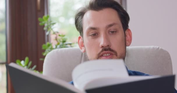 수염을 기른 젊은 남자가 흰 의자에 앉아 책을 읽고 있는 모습을 확대 해 보 세요. 젊은 남자가 집 창문에서 휴식을 취하고 있고 배경에 푸른 식물들이 있습니다. — 비디오