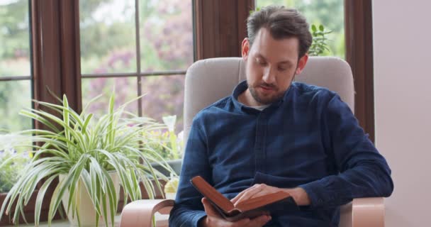 若いですひげを生やした男読書しながら、白い椅子に座って若い男リラックスホームウィンドウと緑の植物を背景に — ストック動画