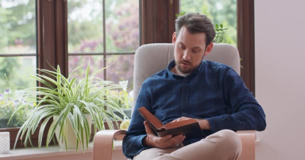 Close up portret młodego brodatego człowieka czytanie książki siedząc na białym krześle z jedną nogą skrzyżowane Młody człowiek relaks w domu Okno i zielone rośliny na tle — Wideo stockowe