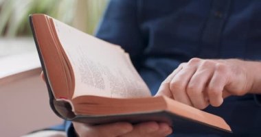 Erkek ellerinin kitap tutuş görüntüsü. Genç adam beyaz sandalyede otururken kitap okuyor. Pencere ve arka planda yeşil bitkiler.