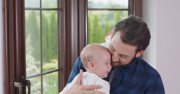 Baard en mosterd vader houdt zijn schattige baby, blijft naast het raam, licht zwaait op de benen, glimlacht, drukt zijn wang op zijn voorhoofd en kust het venster en een witte muur op de — Stockvideo