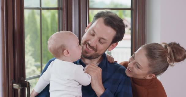 Jóvenes padres felices y su adorable bebé, junto a la ventana El padre barbudo sostiene a su bebé, y baila ligeramente, la madre sonriente está detrás del padre y habla con el bebé Ventana y una pared blanca en la — Vídeo de stock