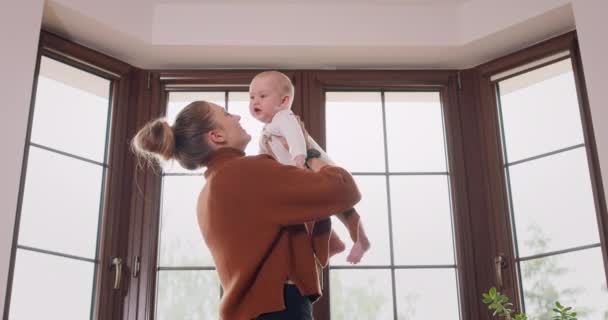 Молодая мать держит на руках своего очаровательного ребенка и остается с ним перед окном, целуя младенца Окно, зеленые растения, и белая стена на заднем плане — стоковое видео