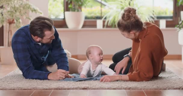 Glimlachend jong koppel liggend samen op tapijt op hun woonkamer vloer thuis met hun schattige baby Baby kijkt naar moeder en katheert haar met zijn handen, baard vader streelt baby op zijn rug — Stockvideo