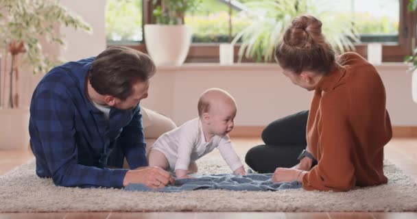 Lachend jong koppel liggend samen op tapijt op hun woonkamer vloer thuis met hun schattige baby Baby kijkt naar moeder en staat op handen en voeten, moeder en bebaarde vader kijken naar baby — Stockvideo