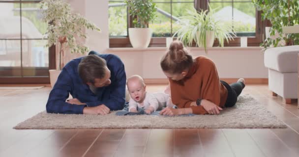 Glimlachend jong koppel liggend samen op tapijt op hun woonkamer vloer thuis met hun schattige baby Baby pakt de deken en staat op handen en voeten, moeder en bebaarde vader kijken naar baby — Stockvideo