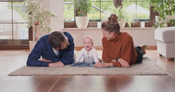 Sorrindo jovem casal deitado juntos no tapete em sua sala de estar chão em casa com seu bebê adorável Bebê sorri e olha para a câmera, mãe e pai barbudo olhar para o bebê e beija-lo — Vídeo de Stock