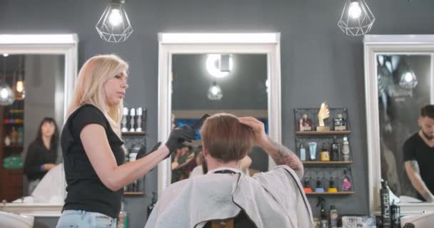 Парикмахерша с боком повернулась к камере, расчесывает волосы молодого бородатого человека, сидящего на парикмахерском стуле спиной к ней, лицом к зеркалу Серая стена с косметикой — стоковое видео