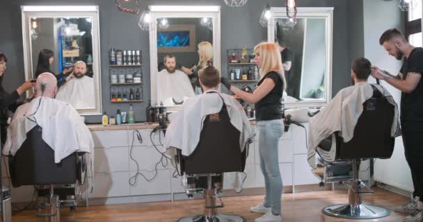 Tři holiči s otočenou stranou na kameru, pracují se svými klienty, kteří sedí na holičských židlích, čelem ke zrcadlu Šedá stěna se zrcadly a kosmetikou na policích v pozadí — Stock video