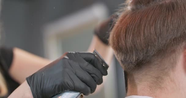 Close-up de mulheres barbeiros mãos, que cortadores com cortador de cabelo e pentear jovens barbudos cabelo, que está sentado na cadeira barbeiros com as costas para ela, de frente para o espelho — Vídeo de Stock