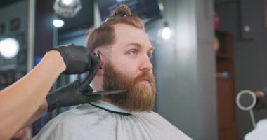 Berber koltuğunda oturan, aynaya bakan genç sakallı adamın yakın yüzü ve kadın berber saçını saç makası ve tarak ile kulağının üzerinden kesiyor ve onunla başka bir berber ile konuşuyor.