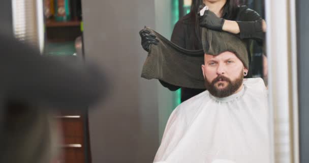 Отражение в зеркале молодого бородатого человека, который сидит на стуле парикмахера, лицом к зеркалу, и женщина парикмахер, который обертывает черное полотенце вокруг мужчин чистой бритые головы парикмахеры руки и — стоковое видео