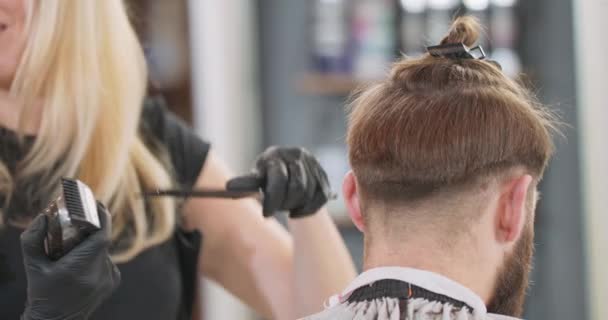 Detailní záběr ženských holičských rukou, které stříhají střihačem vlasů a česají mladé vousaté muže, kteří sedí na holičské židli zády k ní, čelem k zrcadlu. — Stock video