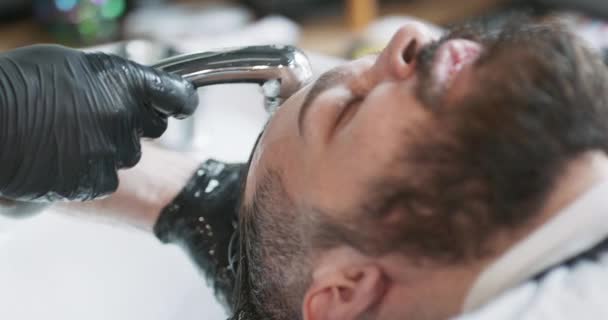 Närbild av ung skäggig man, sitter bredvid tvättställ för att tvätta huvudet, med cape, och tatuerade händer manliga frisör, som häller vatten över håret — Stockvideo