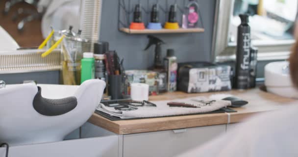 Żeński fryzjer zmienia osprzęt przycinający na szarym ręczniku, gdzie brzytwa, nożyczki do golenia i osprzęt do przycinania są, na szafce, obok umywalki do mycia głowy — Wideo stockowe