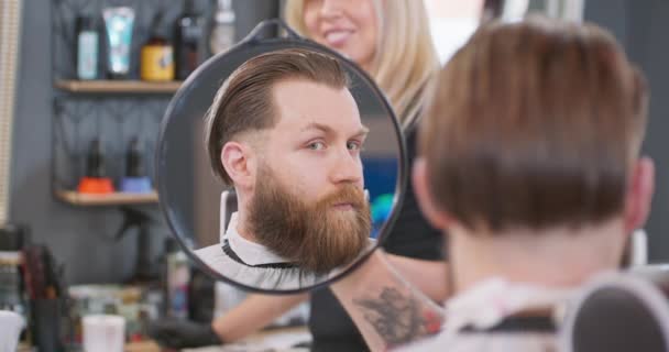 Отражение в зеркале молодого бородатого человека, сидящего на стуле парикмахера, держит зеркало для бритья и оценивает качество работы парикмахеров. — стоковое видео