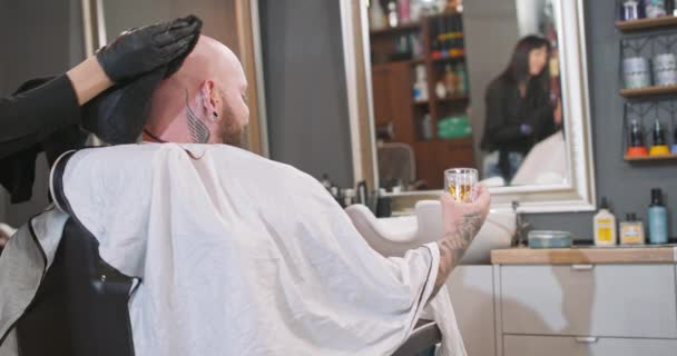 Mladý vousatý muž, s tetováním na krku, který sedí na holičském křesle, s pláštěnkou, drží vytetovanou rukou whisky ve své sklenici, čelem ke zrcadlu, zatímco holič si čistí čistotu — Stock video