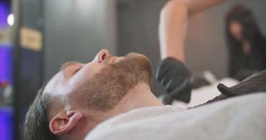 Genç sakallı adamın kafası berber koltuğunda oturuyor, pelerinli, ve kadın berberin elleri, tıraş olmuş boynunu talk pudrasıyla süpürüyor arka planda, hafifçe bulanık, başka bir kadın