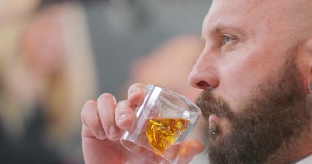 Głowa z bliska młodego brodatego mężczyzny, z pierścieniem podbródkowym, pijącego whisky w szkle w kształcie czaszki, a następnie poprawia wąsy lewą ręką Mężczyzna patrzy przed siebie — Wideo stockowe