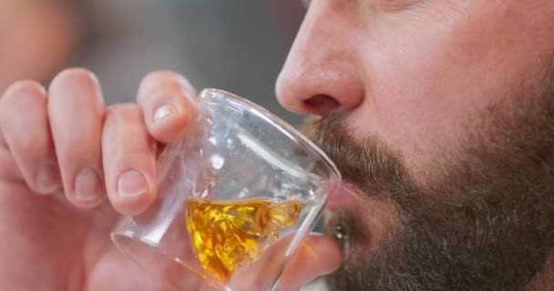 Closeup face of young bearded man, with chin ring, drinking whisky in skull-shaped glass Man is looking straight in front of him Background blurred — Stock Video