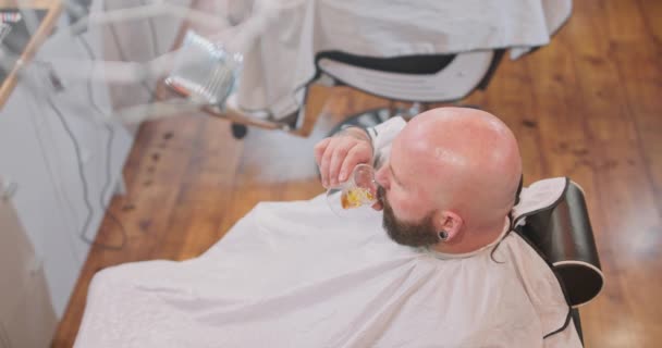 Widok z góry głowy młodych brodatych mężczyzn, z czystą ogoloną głową, siedzi na krześle fryzjerskim, z peleryną, pijąc whisky w kształcie skul szkła, twarzą do podłogi lustra i innym krzesłem fryzjerskim na — Wideo stockowe