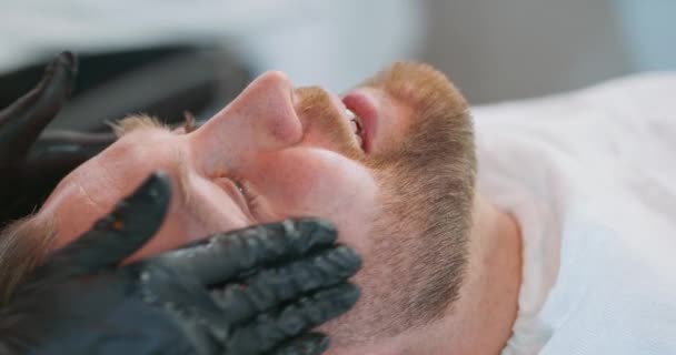 Detailní mladý vousatý muž hlava, na židli holiče, s pláštěm, a ženské holiče ruce v černých rukavicích aplikuje olej a kosmetiku na klienty vousy — Stock video