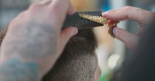 Κοντινό πλάνο τατουάζ άνδρες κουρείς χέρια με ψαλίδι κουρέα και χτένα, κόβει τα μαλλιά του νεαρού άνδρα, ο οποίος κάθεται στην καρέκλα κουρείς — Αρχείο Βίντεο