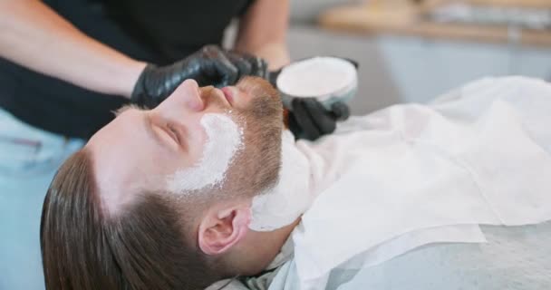 Szoros kéz a női borbély, aki alkalmazni ecsettel borotválkozás krém fiatal szakállas férfiak arc és a nyak — Stock videók