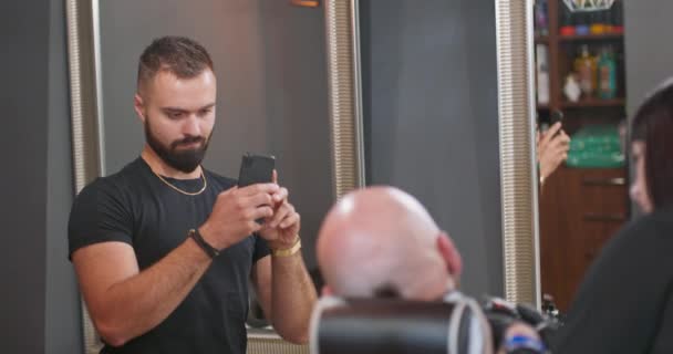 Молодой бородатый мужчина, перед стеной, в черной футболке, со смартфоном в руках, записывает процесс работы парикмахеров, где молодой бородатый мужчина чистая бритая голова сидит на стуле парикмахера — стоковое видео