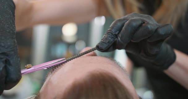 Mâinile strânse ale bărbierului de sex feminin, care taie sprâncenele unui bărbat tânăr cu barbă cu foarfece roz și pieptene — Videoclip de stoc