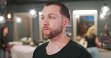 Genç sakallı ve bıyıklı adam, kameranın önünde, berber dükkanında, başını sağa çevirir arka planda, üç berberin müşterilerine hizmet ettiği berber dükkanının arka planında,