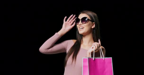 Ein langhaariges brünettes Model mit Sonnenbrille und rosa Einkaufstasche posiert vor schwarzem Studiohintergrund mit Kopierraum — Stockvideo