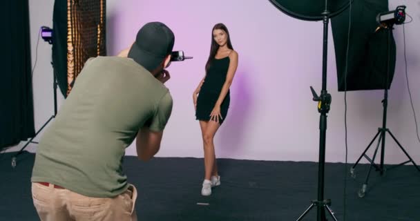 En manlig fotograf med keps tar bilder av en modell mot bakgrund i en studio Fotosession i modern studio — Stockvideo