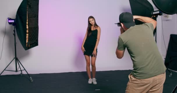 En manlig fotograf med keps tar bilder av en modell mot bakgrund i en studio — Stockvideo