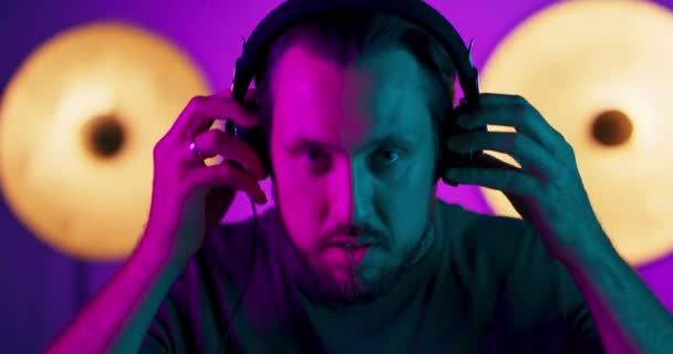 Porträt eines jungen Mannes mit Kopfhörern, während er in die Kamera blickt Männliches Modell in bunten hellen Neonlichtern posiert auf Studiohintergrund — Stockvideo
