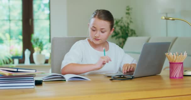 Cute teen girl siedzi przy stole, z laptopem, odrabia pracę domową, pisząc ołówkiem w notatniku Książki, ołówki, okulary i lampa są na stole Sofa, białe ściany, okno w tle — Wideo stockowe