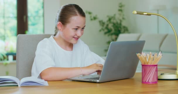 可爱的少女坐在桌旁，拿着笔记本电脑，写着，笑着写着便条和铅笔，桌上放着一盏灯。绿色的植物，沙发，白色的墙壁，背景窗 — 图库视频影像