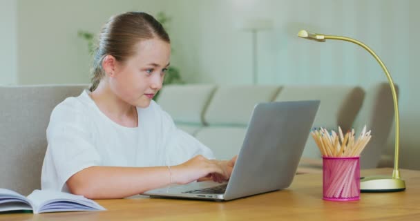 Linda chica adolescente se sienta en la mesa, con el ordenador portátil, y escribe, sonriendo Notas y lápices, y una lámpara están en la mesa Plantas verdes, sofá, paredes blancas en el backgroung — Vídeo de stock