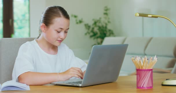 Cute teen girl sits at the desk, with laptop, and writes, smiling Notes and pencils, and a lamp are on the table Green plants, sofa, white walls and a window at backgroung — Stock Video