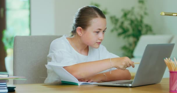 Linda chica adolescente en el escritorio, con el ordenador portátil, haciendo los deberes, comprobar las tareas en el ordenador portátil, escritura en cuaderno Libros, lápices, lámpara están en el escritorio Plantas verdes, sofá, paredes blancas y ventana en backgroung — Vídeos de Stock